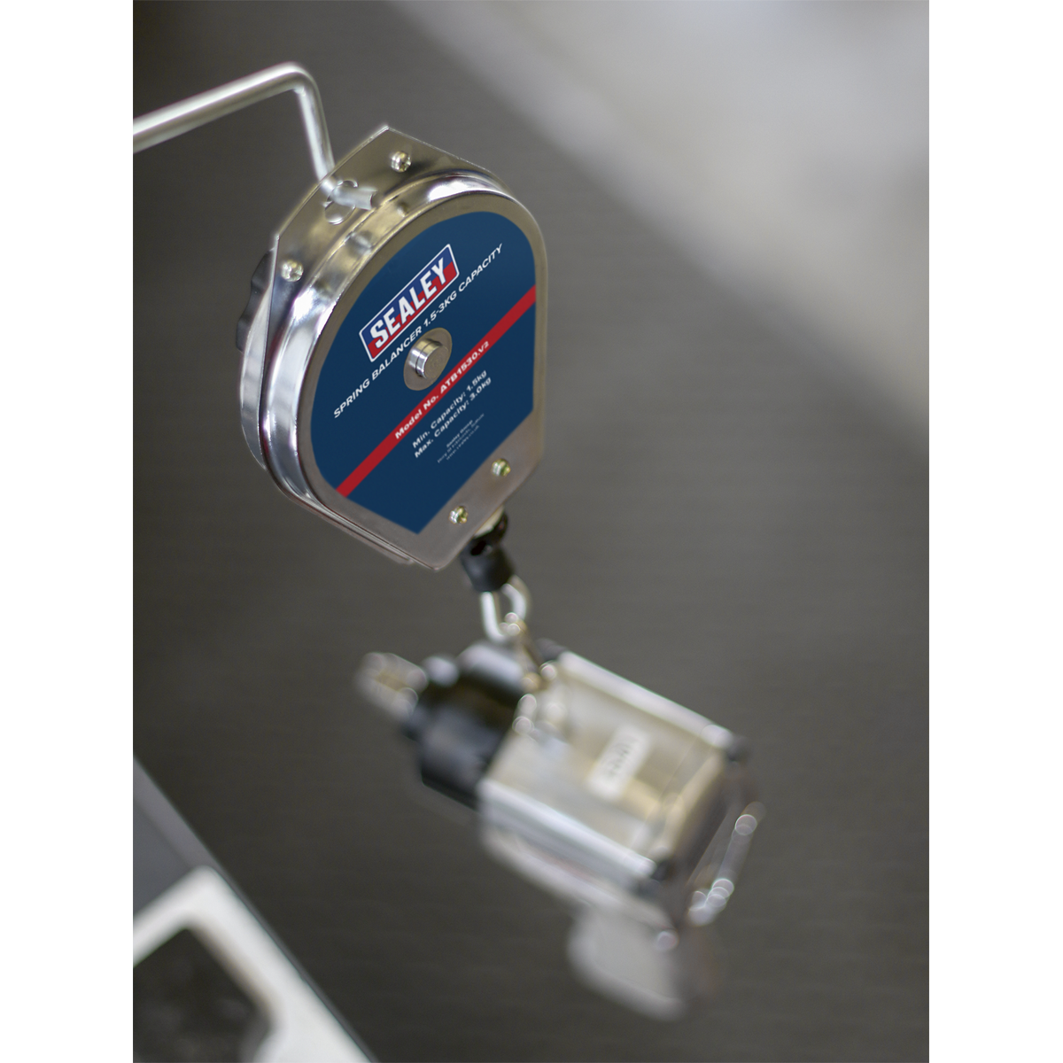 Close-up of a Sealey Spring Balancer 1.5-3kg Capacity - ATB1530 displaying the brand logo, with an attached pneumatic tool hanging below for weighing. The tool support system minimizes operator fatigue, and the background is blurred.