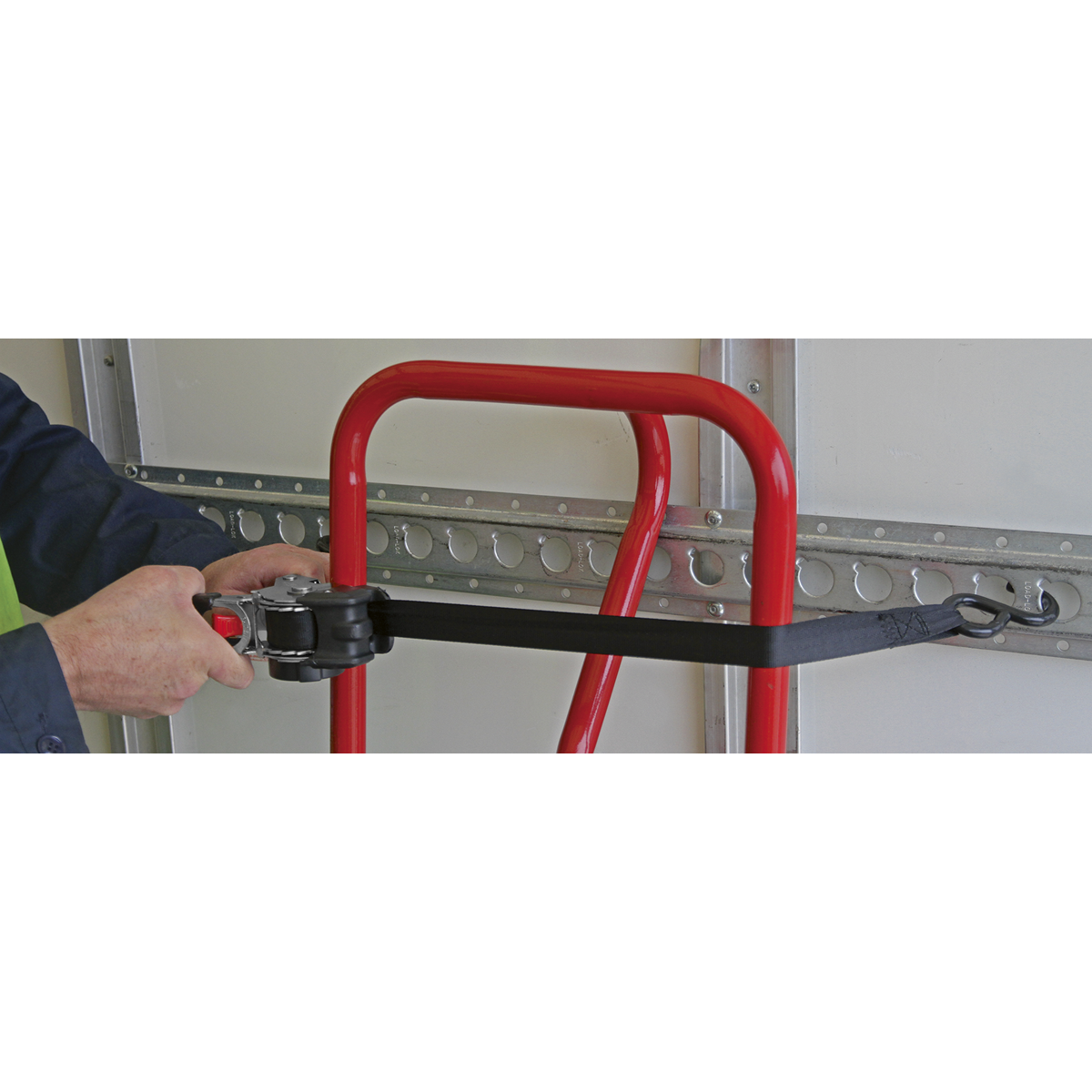 Using the Sealey Auto Retractable Ratchet Tie Down 25mm x 3m - ATD25301, a person secures a black cargo strap to a rail on a red hand truck.