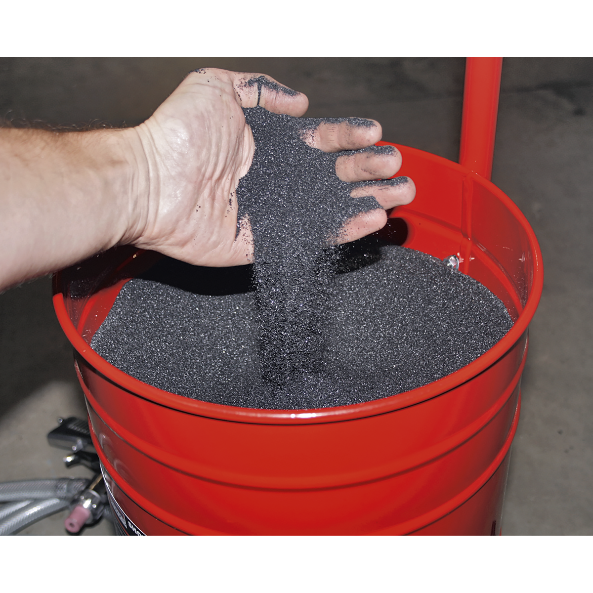 A handheld shot blaster is being operated, with a hand carefully pouring Sealey Shot Blasting Grit from a 25kg bag (B/25KG) into a red container. The container, likely used in a workshop, resembles a barrel or bucket.