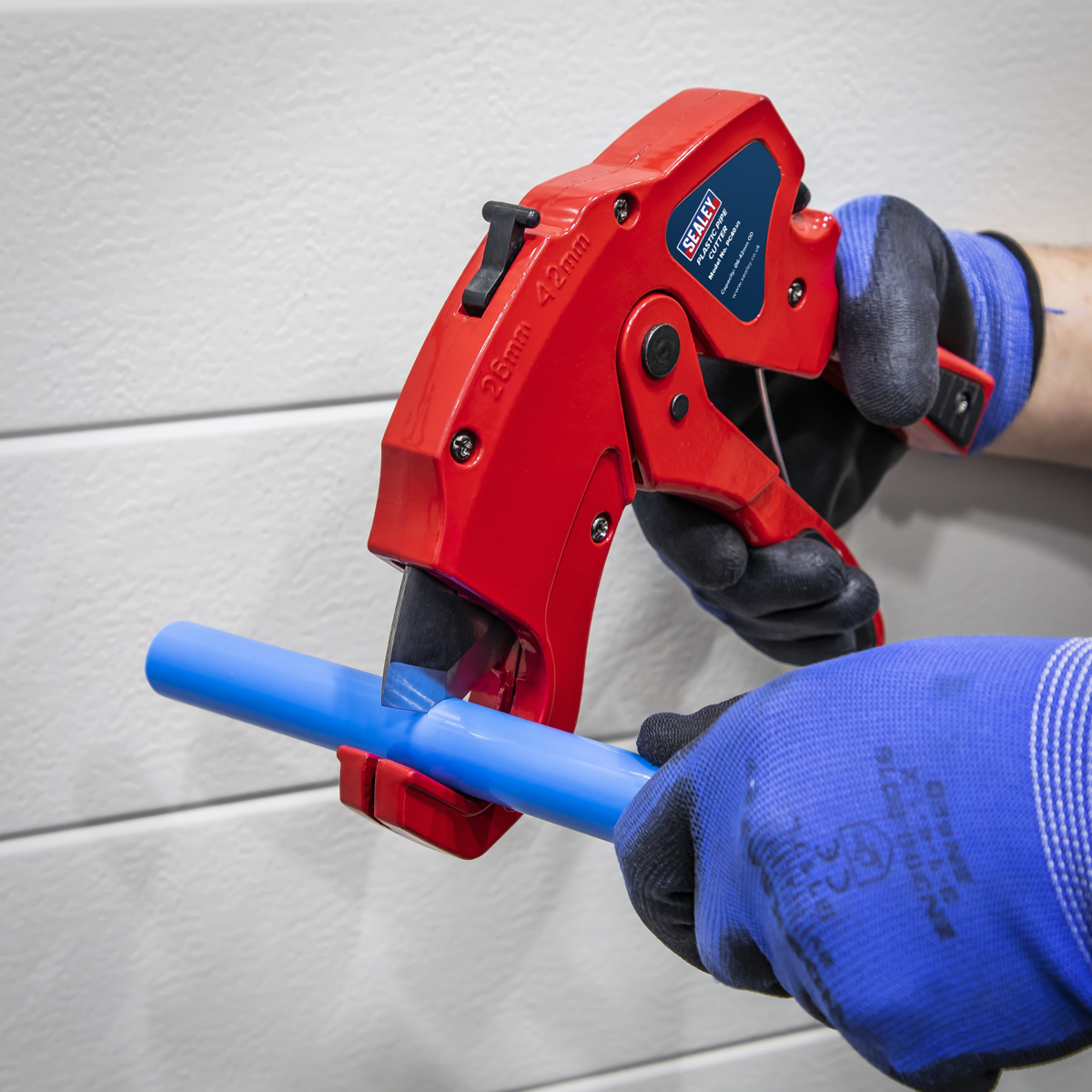 A person wearing blue gloves uses a Sealey John Guest Speedfit red pipe cutter to cut through a blue Rigid Nylon Pipe 28mm x 3m (Pack of 5 - CAS28NP) against a white, textured wall.