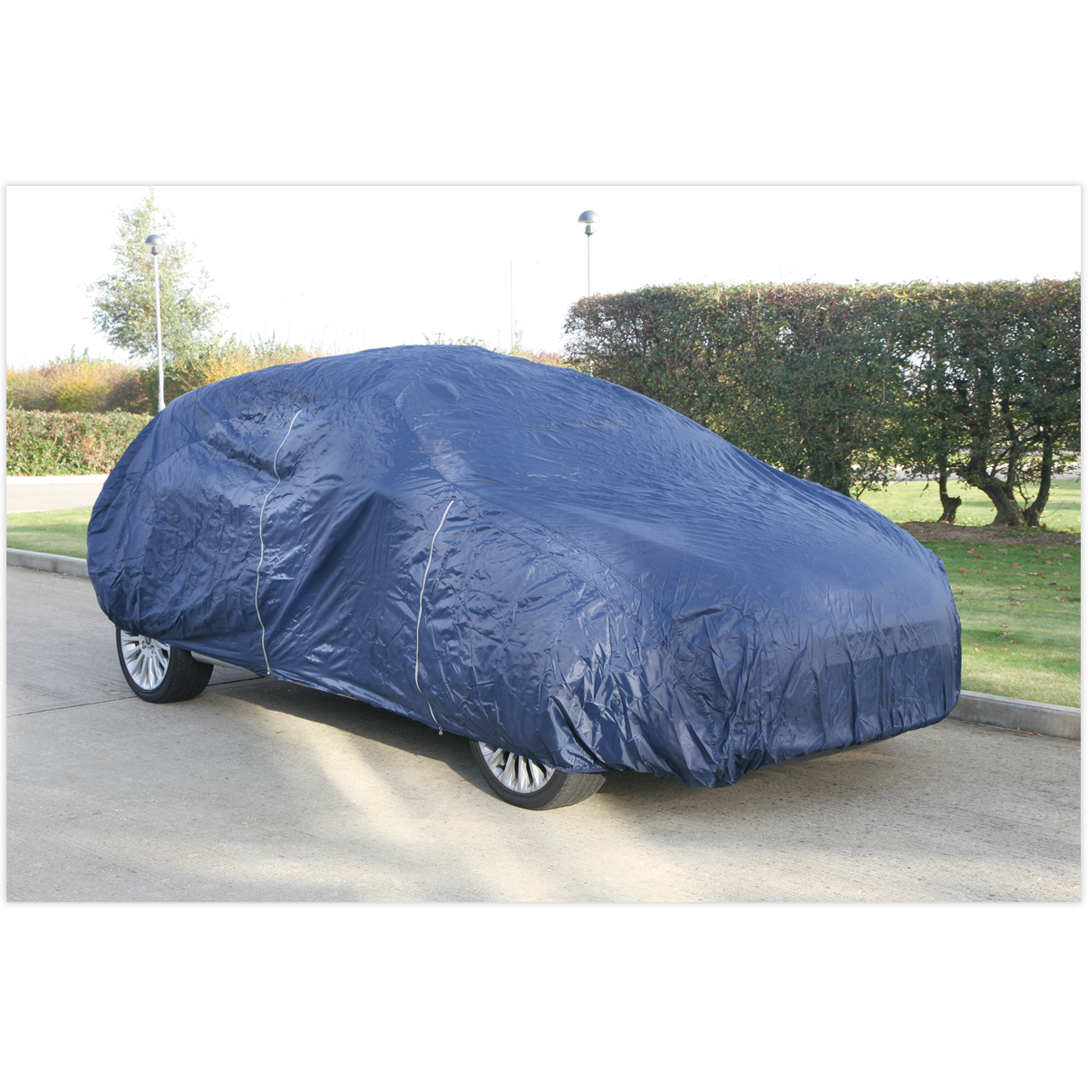 A parked car is completely covered with the Sealey Car Cover Lightweight Large 4300 x 1690 x 1220mm - CCEL, made of lightweight material on a street lined with greenery and a hedge in the background, ensuring dust protection.