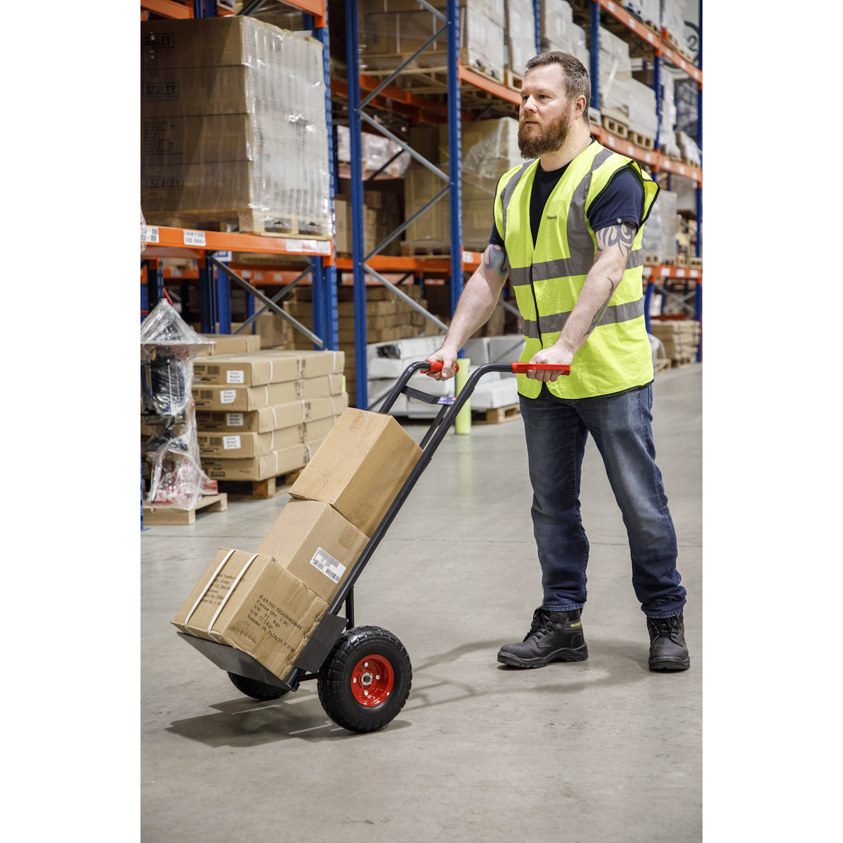 A person wearing a high-visibility vest uses the Sealey Heavy-Duty Sack Truck with PU Tyres 250kg Capacity (CST986HD) to move boxes in a warehouse.