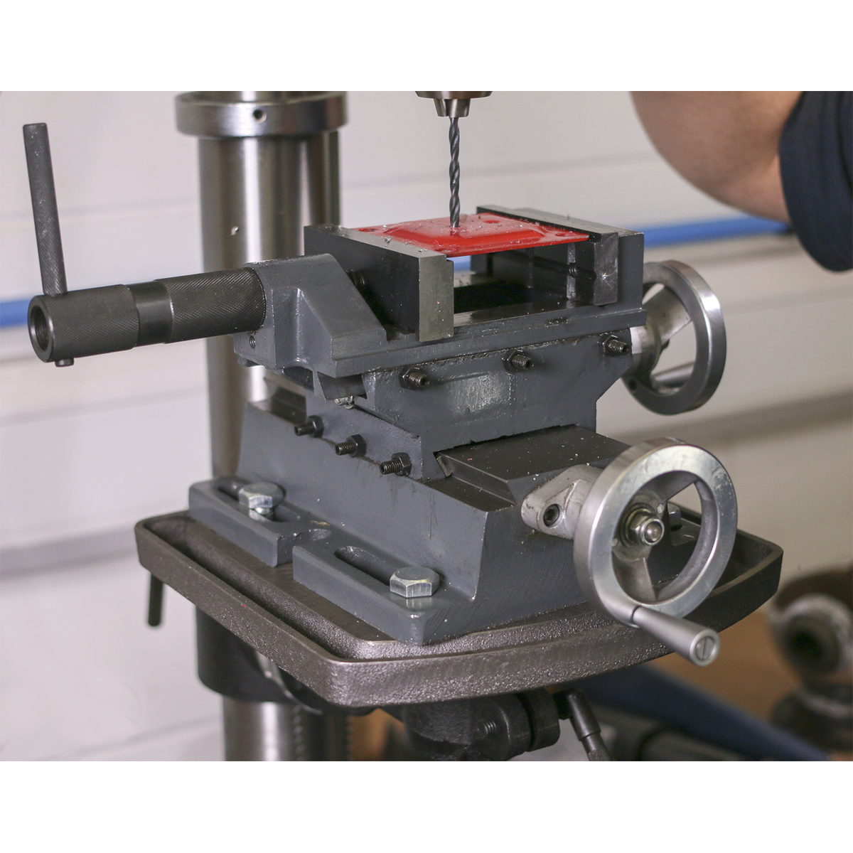 A close-up of a milling machine in use, showing the drill bit and metal workpiece being securely held by the Sealey Cross Vice 100mm Professional - CV4P during a drilling operation.