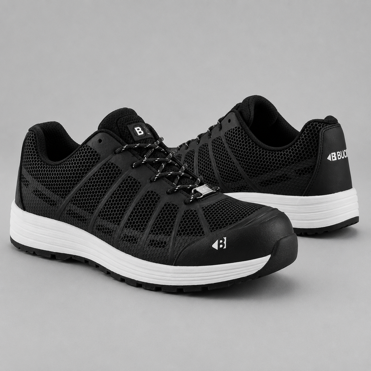 A pair of black protective work shoes with white midsoles, shown from the side and back on a neutral gray background.