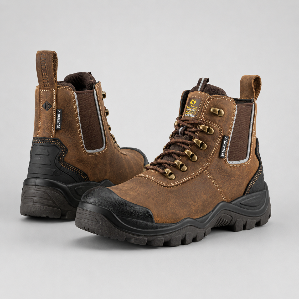 A pair of brown leather work boots with black rubber toe caps and soles, featuring elastic side panels and gold-tone metal eyelets, photographed on a light gray background.