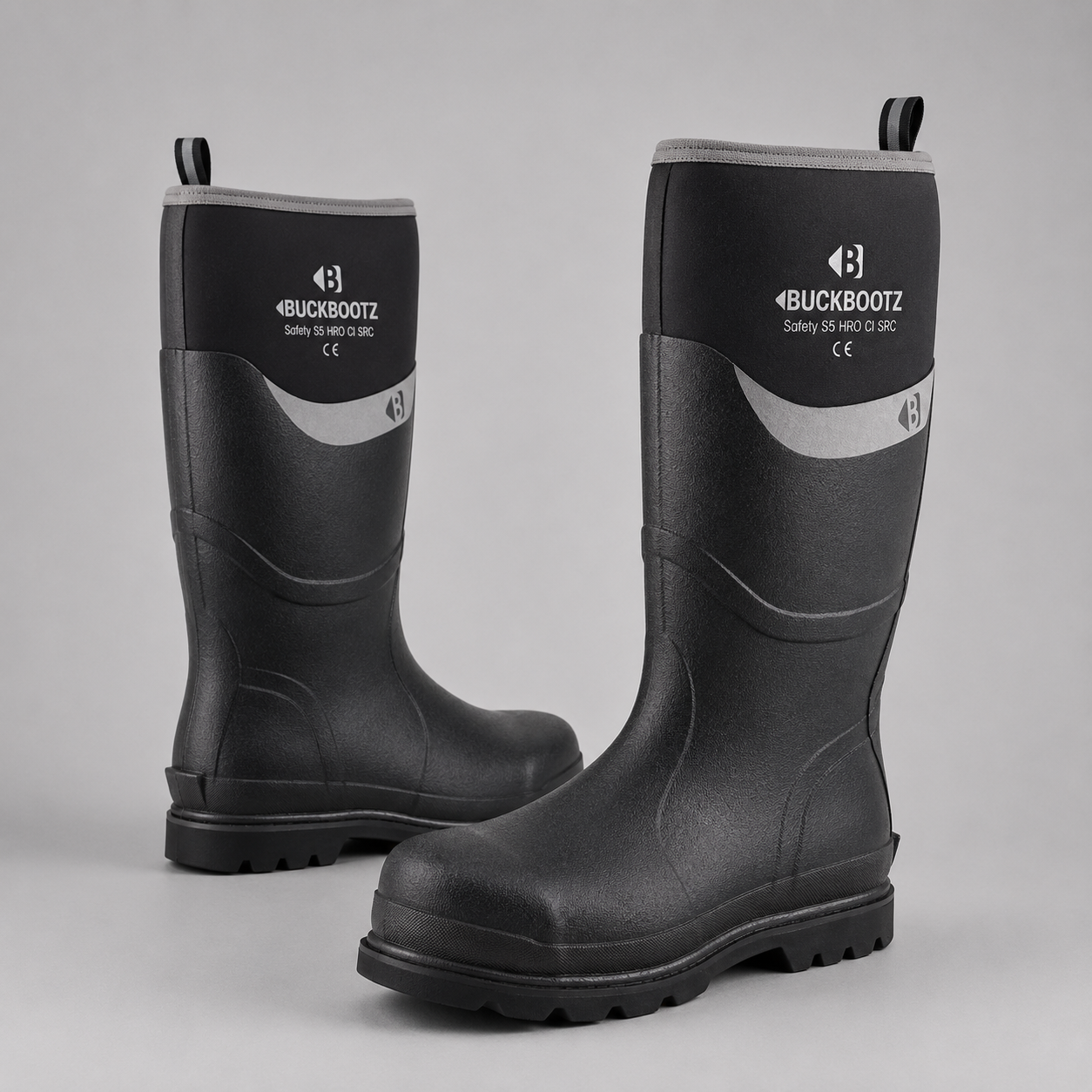 A pair of Buckbootz safety work boots in black rubber with a gray curved accent stripe and white logo branding on the upper shaft, set against a neutral gray background.