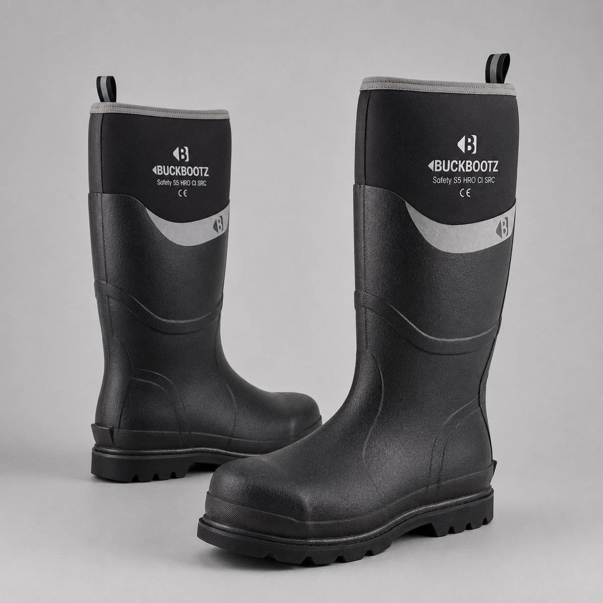 A pair of Buckbootz safety work boots in black rubber with a gray curved accent stripe and white logo branding on the upper shaft, set against a neutral gray background.