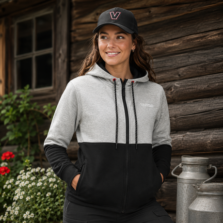 A woman wearing a color-block zip-up hoodie with a light gray upper section and black lower section, paired with a black baseball cap, standing in front of a rustic wooden cabin.