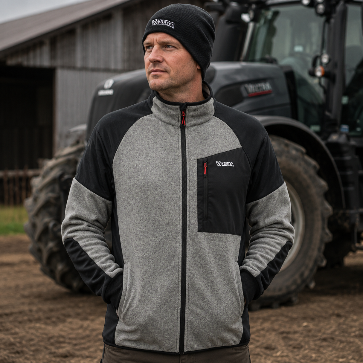 A man wearing a gray and black color-blocked fleece jacket and a black knit beanie with 'Volkswagen' printed on it, standing in front of a tractor on a farm.