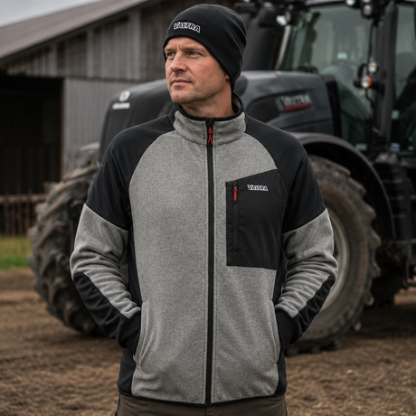 A man wearing a gray and black color-blocked fleece jacket and a black knit beanie with 'Volkswagen' printed on it, standing in front of a tractor on a farm.