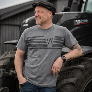 A man wearing a heather gray Valtra t-shirt with a dark gray striped graphic and 'V' logo, standing in front of a black Valtra tractor.