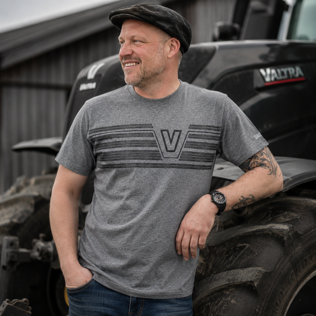 A man wearing a heather gray Valtra t-shirt with a dark gray striped graphic and 'V' logo, standing in front of a black Valtra tractor.