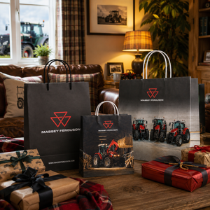 Three Massey Ferguson branded shopping bags and a small branded box are arranged on a wooden table in a cozy living room, surrounded by wrapped Christmas gifts. The bags feature the Massey Ferguson logo and tractor imagery, with a window, sofa, lamp, and plants visible in the background.