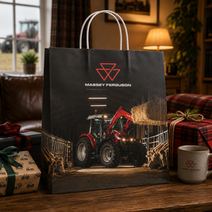 A black Massey Ferguson shopping bag printed with a red tractor and barn scene, standing on a wooden table in a cozy living room with wrapped gifts and a mug nearby.