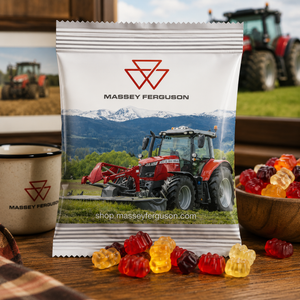 A bag of Massey Ferguson branded gummy candies sits on a wooden table, with red and yellow gummies scattered in front and a bowl of more gummies to the right. A tractor is pictured on the bag, and a blurred real farm setting with a tractor appears in the background.