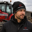 A man wearing a black knit beanie and black zip-up jacket with red and white Massey Ferguson logos, standing in front of a red tractor on an outdoor farm setting.