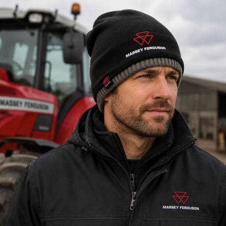 A man wearing a black knit beanie and black zip-up jacket with red and white Massey Ferguson logos, standing in front of a red tractor on an outdoor farm setting.