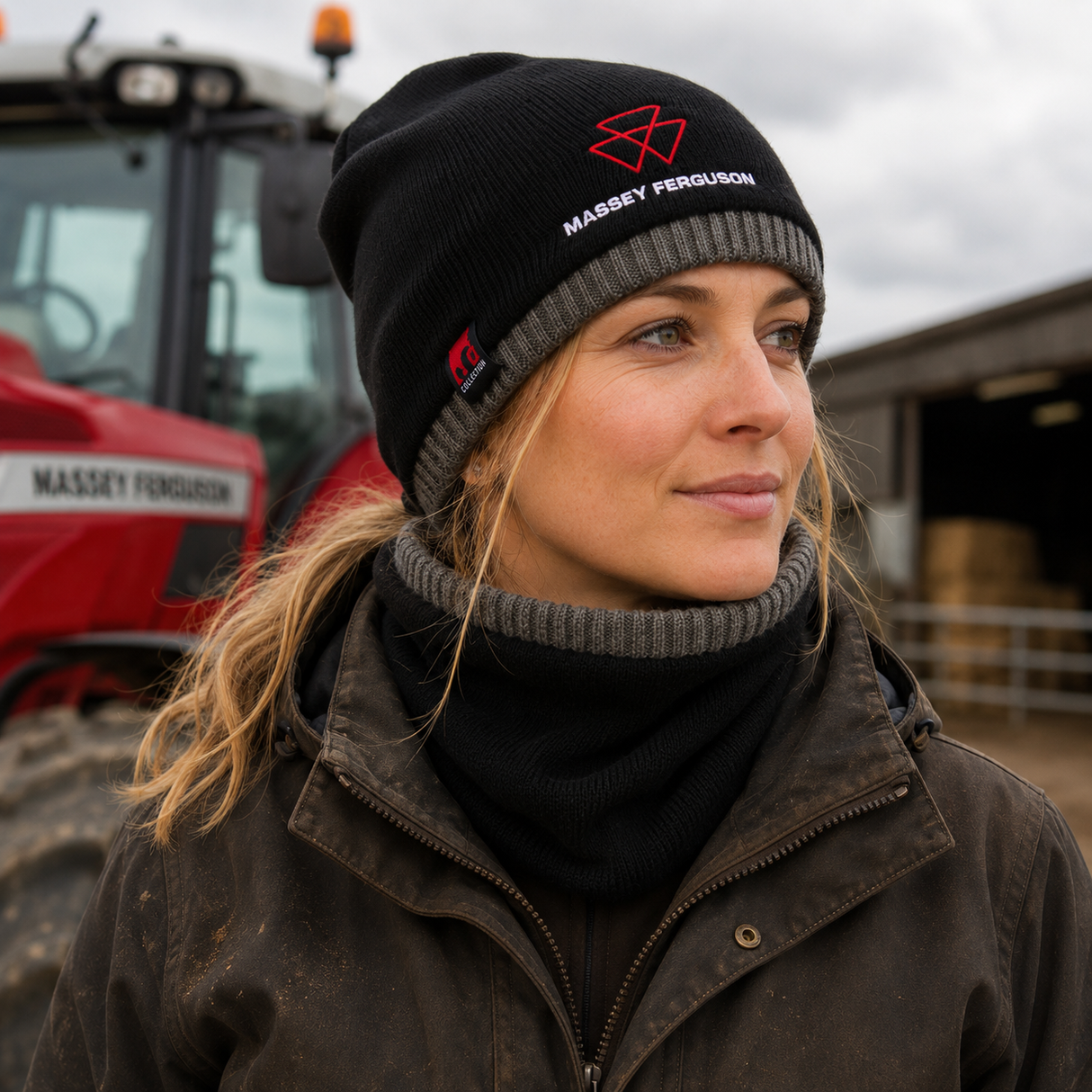 A woman wearing a black knit beanie with a red Massey Ferguson logo and white text, standing outdoors in front of a red tractor and barn under an overcast sky.