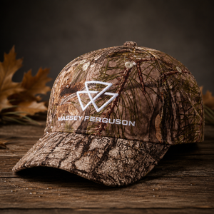 A camouflage baseball cap with a white embroidered Massey Ferguson logo and text on the front, displayed on a rustic wooden surface with autumn leaves in the background.
