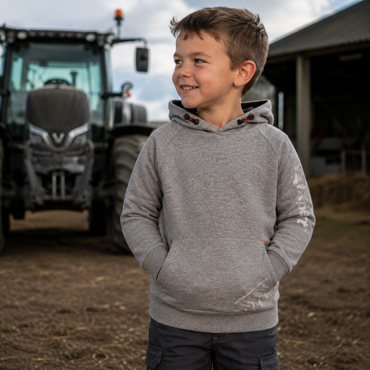 A young boy wearing a heather gray pullover hoodie with a kangaroo pocket stands on a farm in front of a tractor and barn.
