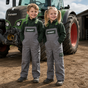 Two children standing in front of a green Fendt tractor on dirt ground, wearing matching gray and dark green work overalls with FENDT logos on the chest.