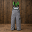 Gray Fendt bib overalls with green suspenders and a green chest patch displaying the FENDT logo, shown against a dark wood plank background.