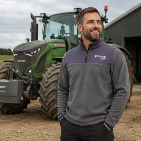 A smiling man stands outdoors in front of a green Vendy tractor and barn, wearing a gray and dark purple quarter-zip fleece pullover with a white Vendy Foli logo on the chest.