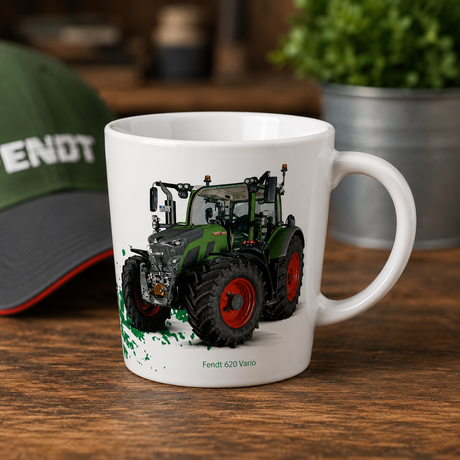 A white ceramic mug featuring a printed illustration of a green and black Fendt 620 Vario tractor with red wheels, displayed on a wooden table with a blurred green cap and plant in the background.