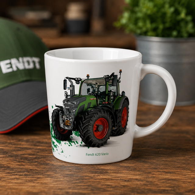 A white ceramic mug featuring a printed illustration of a green and black Fendt 620 Vario tractor with red wheels, displayed on a wooden table with a blurred green cap and plant in the background.