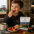 A young boy in a black Massey Ferguson hoodie sits at a wooden table, eating red gummy candies from a bowl while a packaged bag of Massey Ferguson gummies sits next to him. The scene is set in a rustic barn with a tractor in the background.