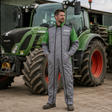 A man wearing a gray and green agricultural jumpsuit stands in front of a green VendiTech tractor on a farm. The jumpsuit is primarily gray with green sleeves and shoulder panels, and black zippers and piping details.