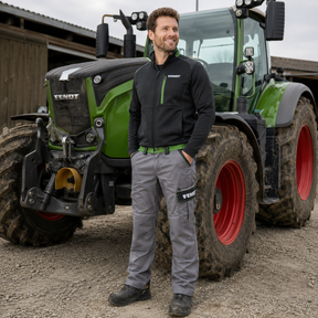 A man wearing a black and green Fenix jacket and gray trousers stands in front of a green and black Fendt tractor on a farm.