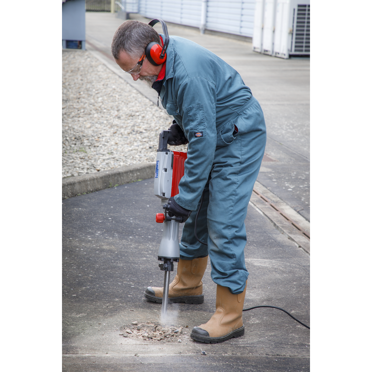 A person in blue coveralls and protective gear is using the Sealey Demolition Breaker Hammer 1600W/230V - DHB1600 on a concrete surface outdoors.