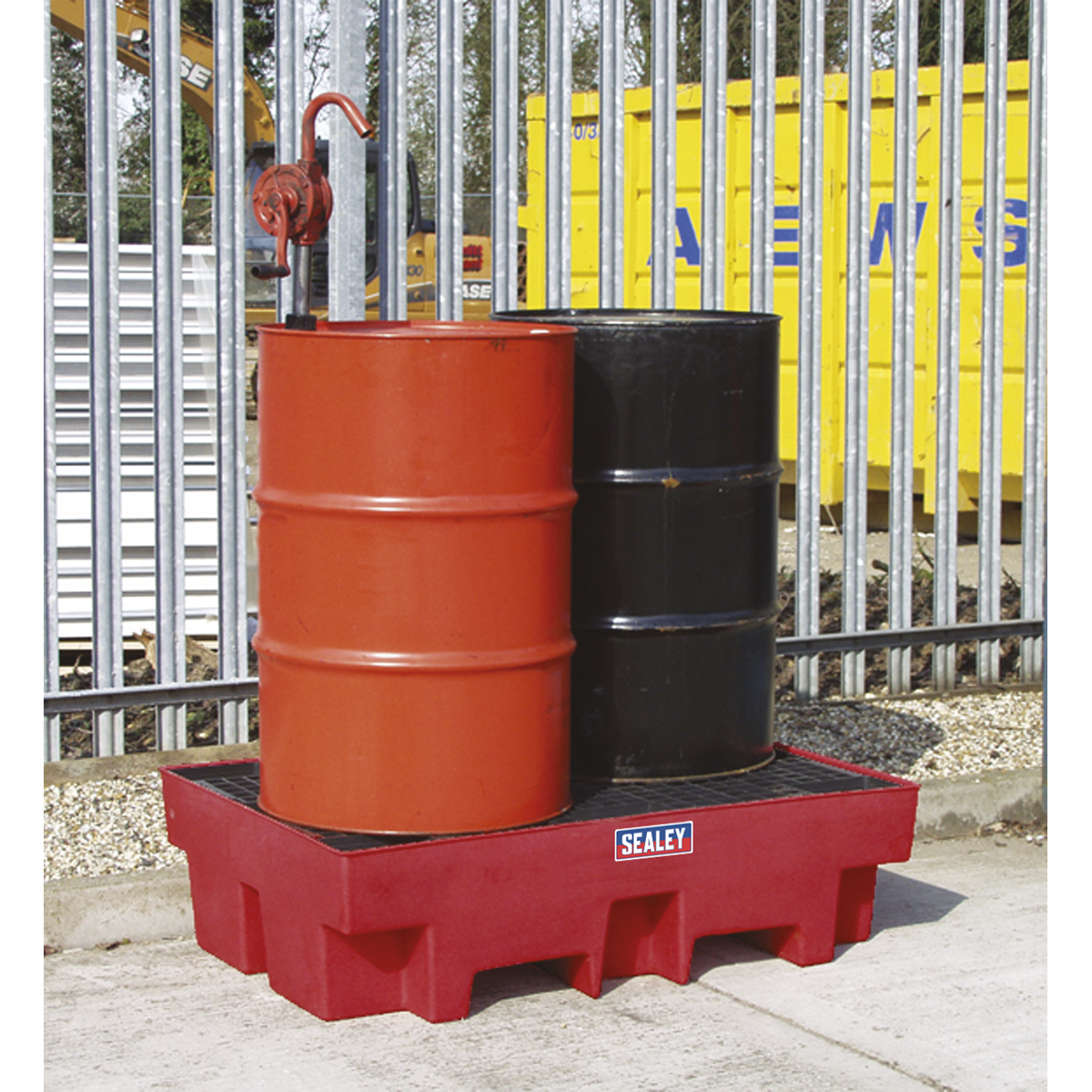 Two industrial barrels, one red and one black, are placed on a Sealey Barrel Bund Polyethylene 1220 x 820 x 330mm - DRP12 spill containment pallet. The red barrel is equipped with a pump. This setup is situated outdoors near a metal fence and yellow container, demonstrating an emphasis on compliance with oil storage regulations.