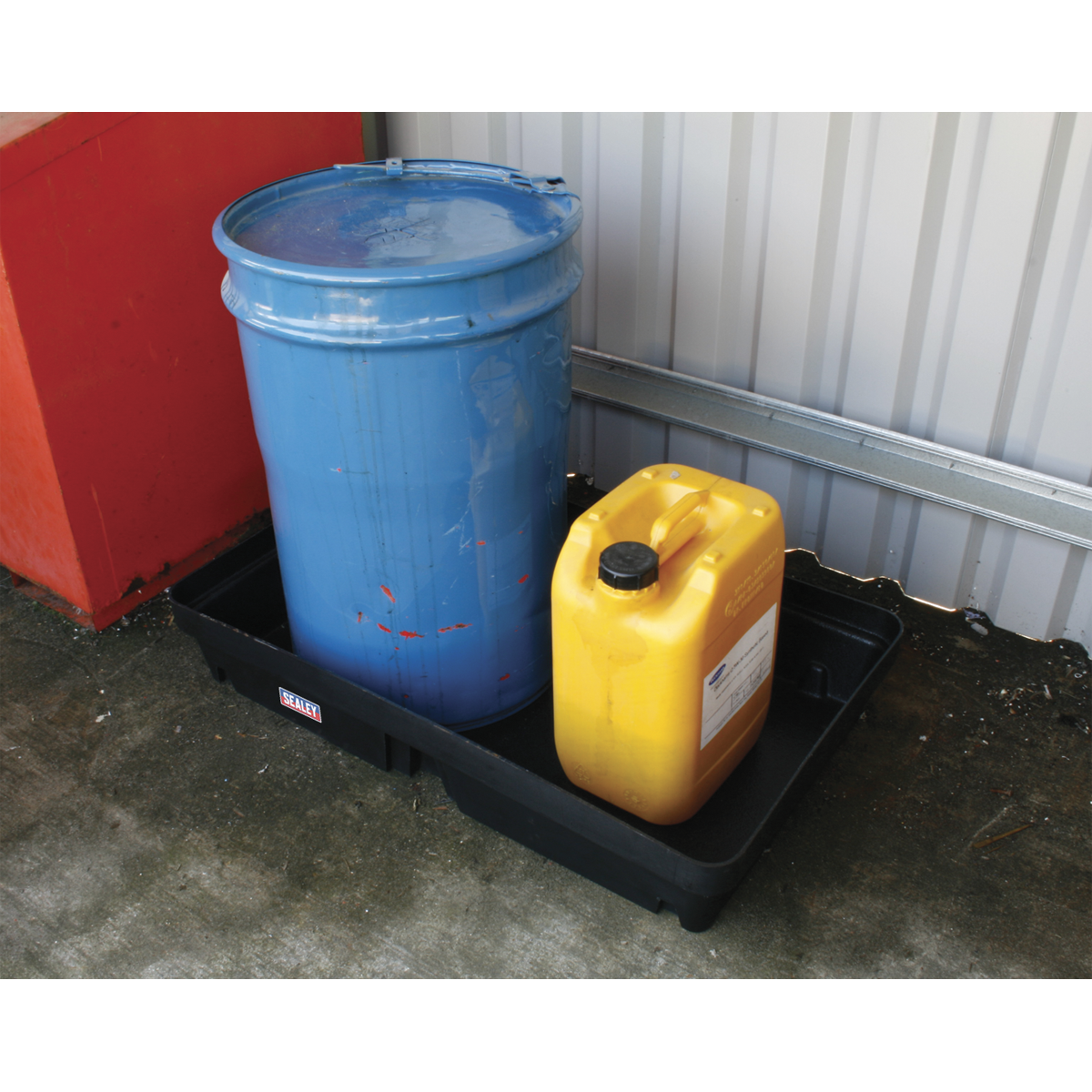 A large blue barrel and a small yellow container sit in a Sealey Spill Tray 60L - DRP32 made of high-density polyethylene against a corrugated metal wall.