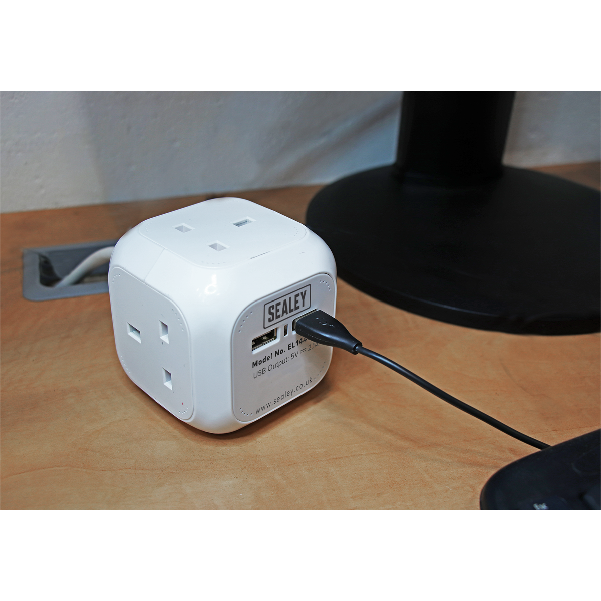 A white Sealey Extension Cable Cube (EL144USB) with its compact, space-saving design sits on a desk, featuring four 230V power outlets and two USB sockets, with a USB cable plugged into one of its ports.