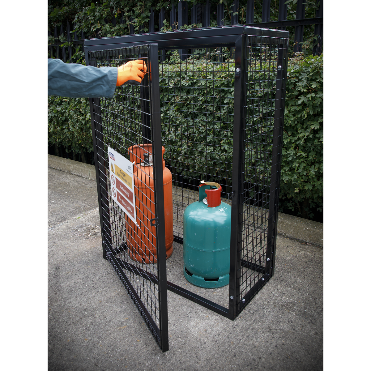 A person opening a Sealey Safety Cage (model GCSC319) made of black, powder-coated galvanised wire mesh containing an orange and a green propane and butane cylinder, with a warning sign posted on the cage door.