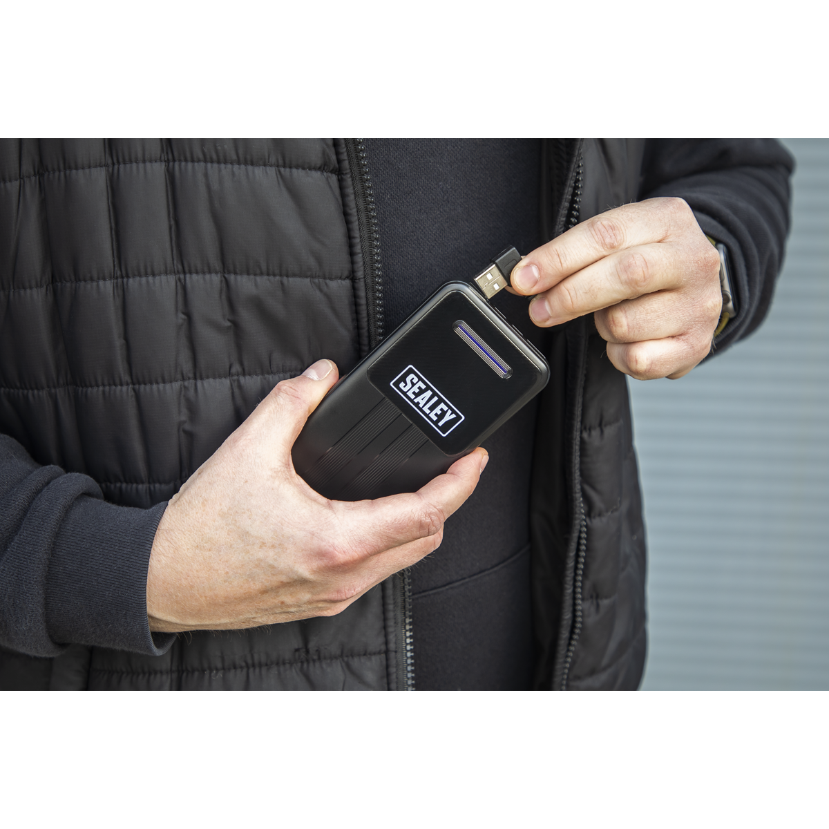 A person plugs a USB cable into a Sealey-branded electronic device while wearing a black 5V Heated Puffy Gilet with three heating zones, standing against a corrugated metal background.