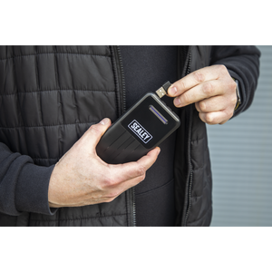 A person plugs a USB cable into a Sealey-branded electronic device while wearing a black 5V Heated Puffy Gilet with three heating zones, standing against a corrugated metal background.