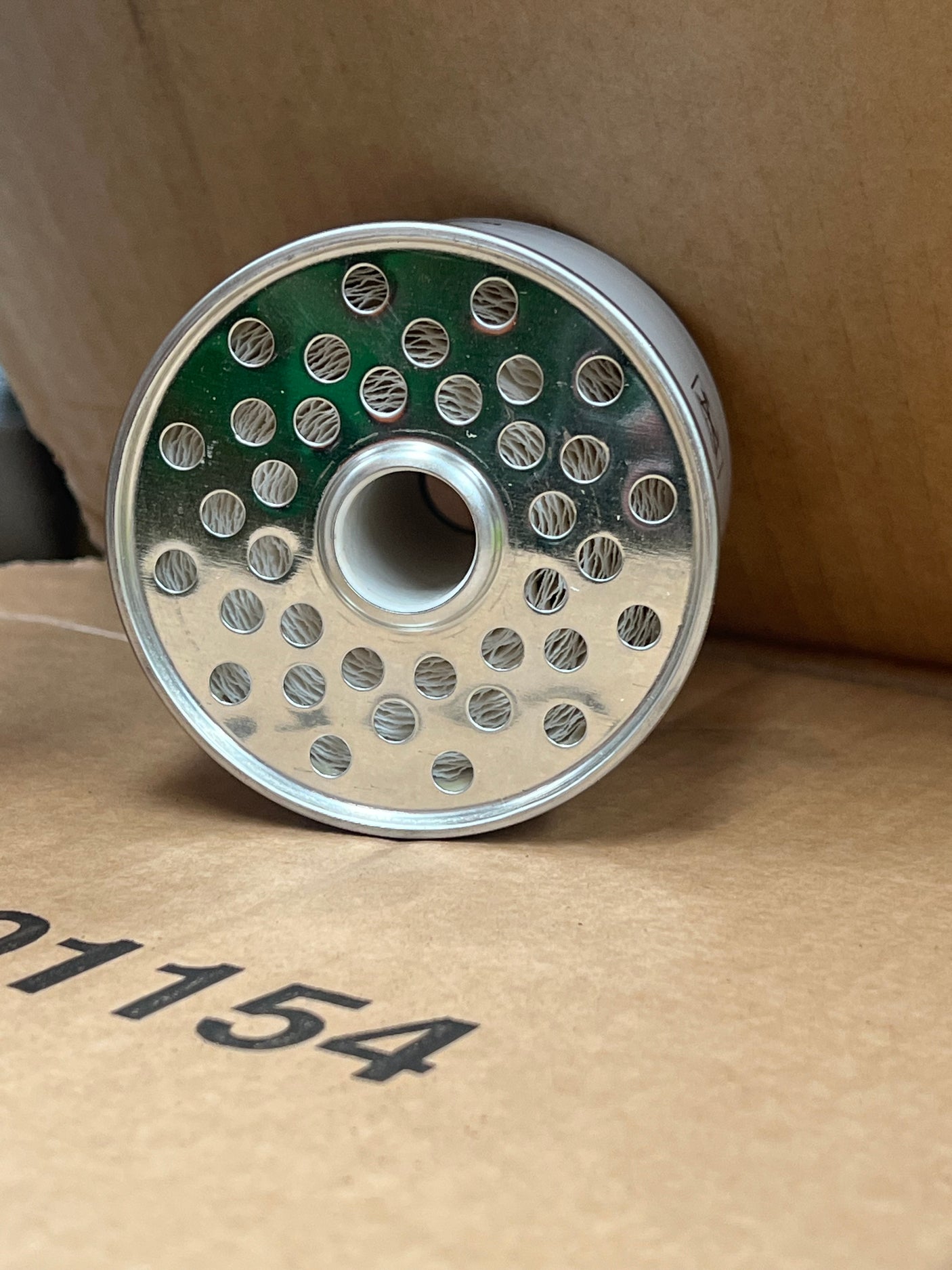 A cylindrical metal component with multiple holes on one end, resembling the Sparex Fuel Filter - Element - FF167A (Sparex Part No. S.109023), is carefully placed on a cardboard surface.