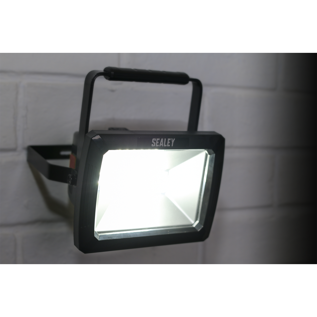 A black Sealey Rechargeable Portable Floodlight 10W SMD LED - LED084 is mounted on a white brick wall, its SMD LED technology illuminating brightly.