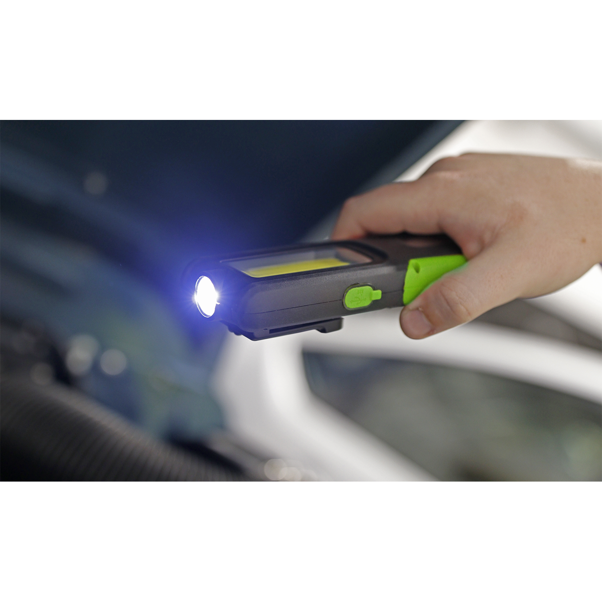 A hand holds a Sealey Rechargeable Inspection Light 5W COB & 3W SMD LED with Power Bank in green (model LED318G), featuring a super bright 5W COB LED light and an illuminated white 3W SMD LED bulb. The background reveals a car with an open hood.