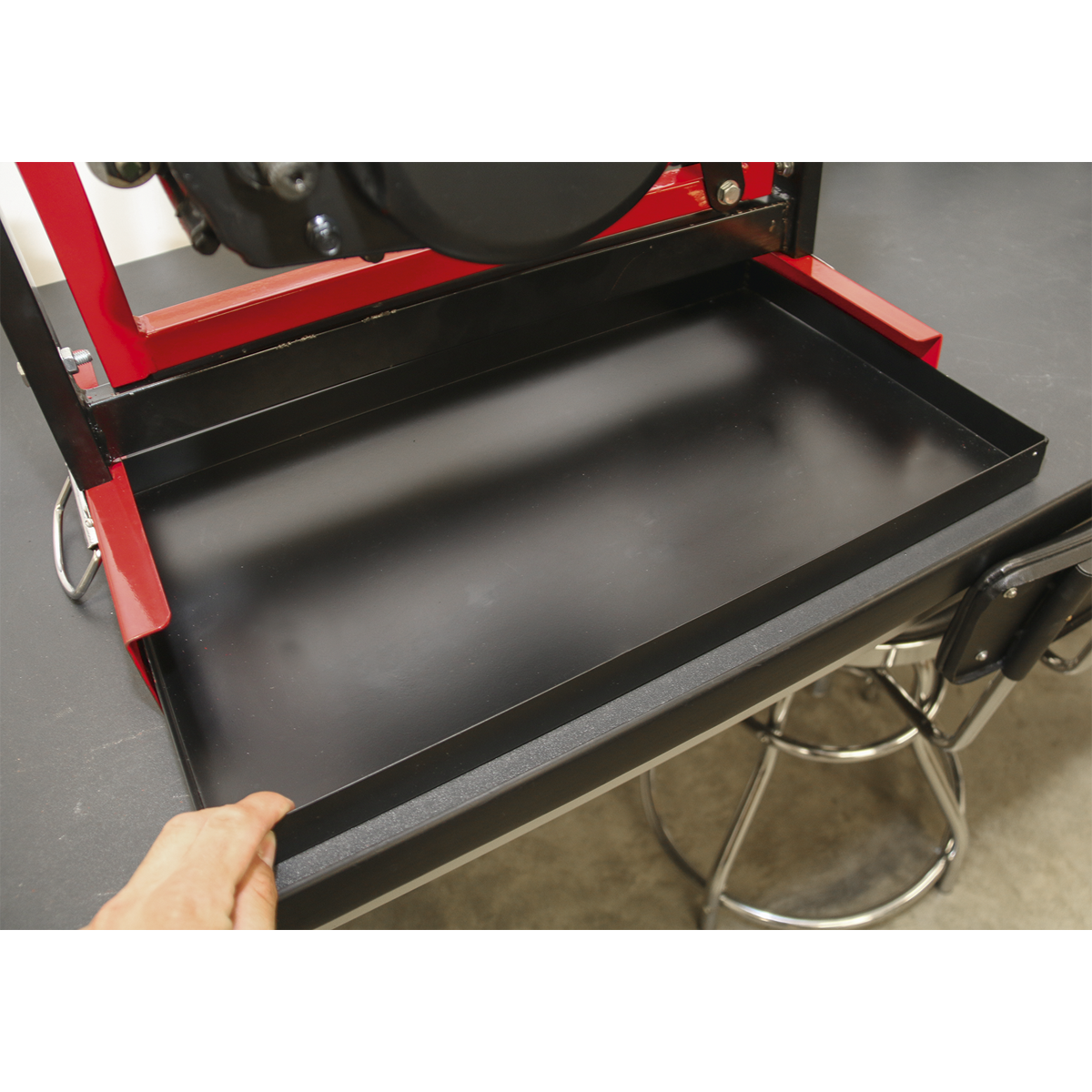 A person pulls a black metal utility tray from a red and black Motorcycle Engine Stand - Single/Twin Cylinder - MES01 by Sealey on a workshop table. A stool is partially visible beside the table, hinting at work involving twin cylinder engines or perhaps some single cylinder engines.