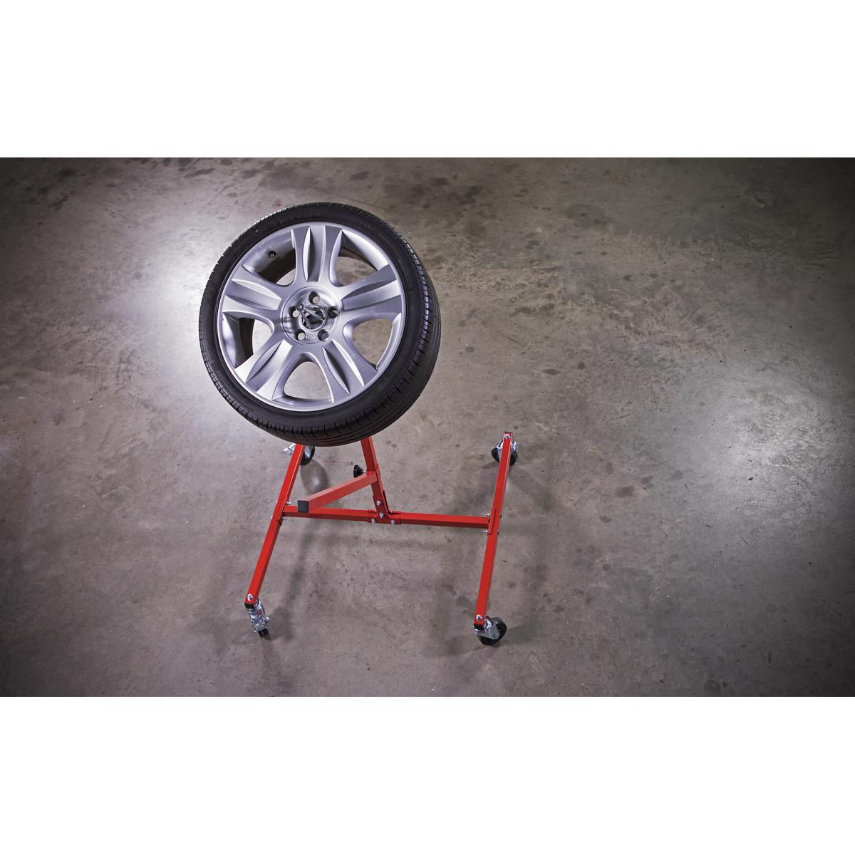 The Sealey MK72 Alloy Wheel Painting/Repair Stand, capable of accommodating wheels up to 22", is mounted on a sturdy red metal frame with adjustable supporting bars and casters, positioned on a concrete floor.