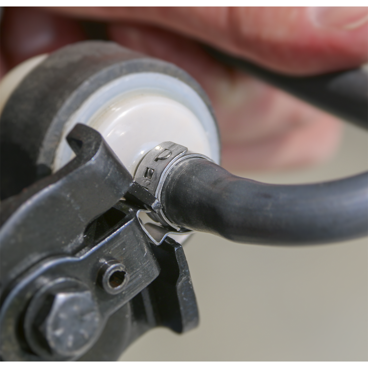 Close-up of a Sealey O-Clip Single Ear Ø13.2-15.7mm (OCSE1315) stainless steel clamp securing a black hydraulic hose onto a white plastic connector, with a hand in the background adjusting the setup.