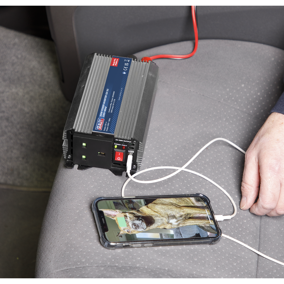 A Sealey Power Inverter 300W 12V DC - 230V~50Hz - PI300 is placed on a car seat, with a smartphone displaying a cat photo connected and charging via the USB port.