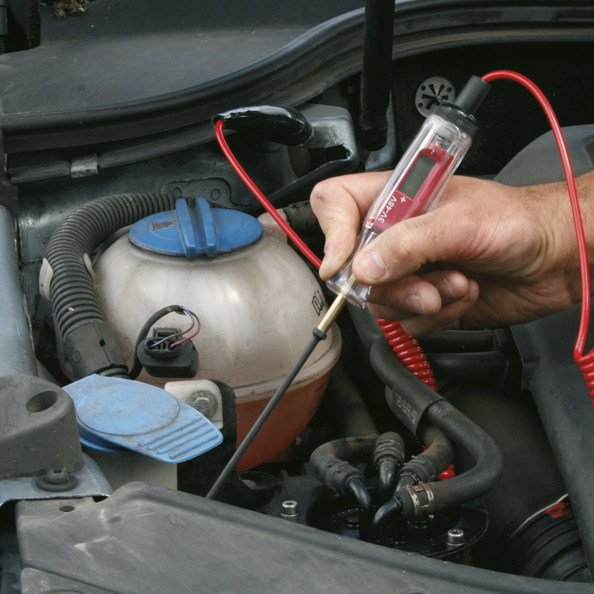 A person uses the Sealey Integrated Test Light/Voltmeter 3-48V - PP5 on a car engine. The tester, equipped with interchangeable probes and an LCD meter, is connected with a red wire. A coolant reservoir with a blue cap is also visible in the background.