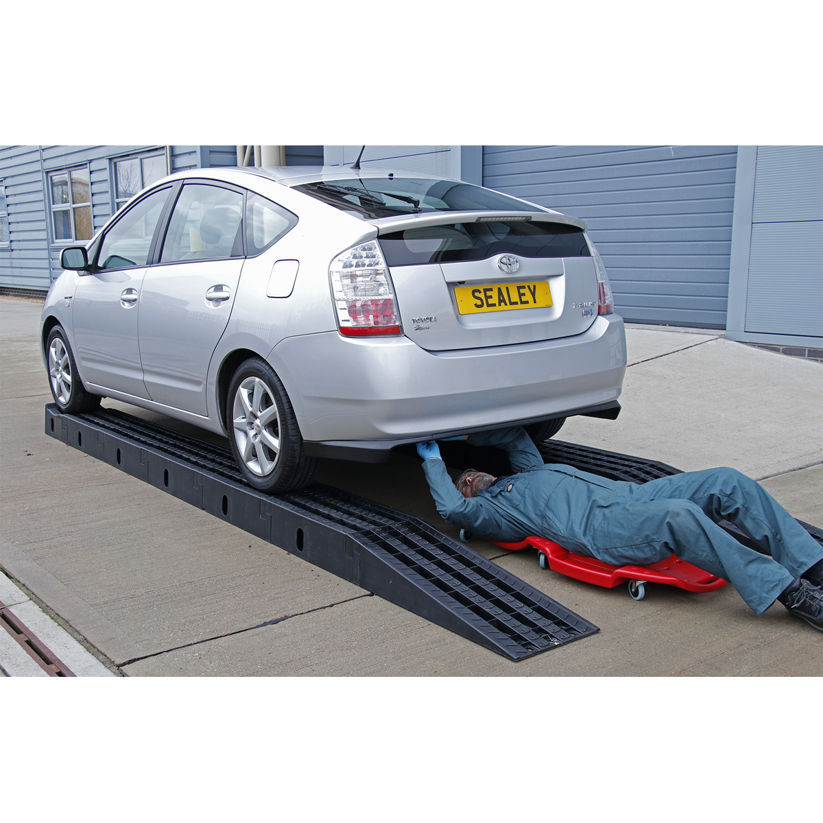 A technician in a blue coverall is lying on a creeper under a silver car, which is raised on Sealey's Modular Pit Ramp Car 4 Tonne - PRK01, boasting a sturdy 4-tonne capacity, in an outdoor setting.