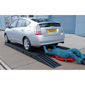 A technician in a blue coverall is lying on a creeper under a silver car, which is raised on Sealey's Modular Pit Ramp Car 4 Tonne - PRK01, boasting a sturdy 4-tonne capacity, in an outdoor setting.