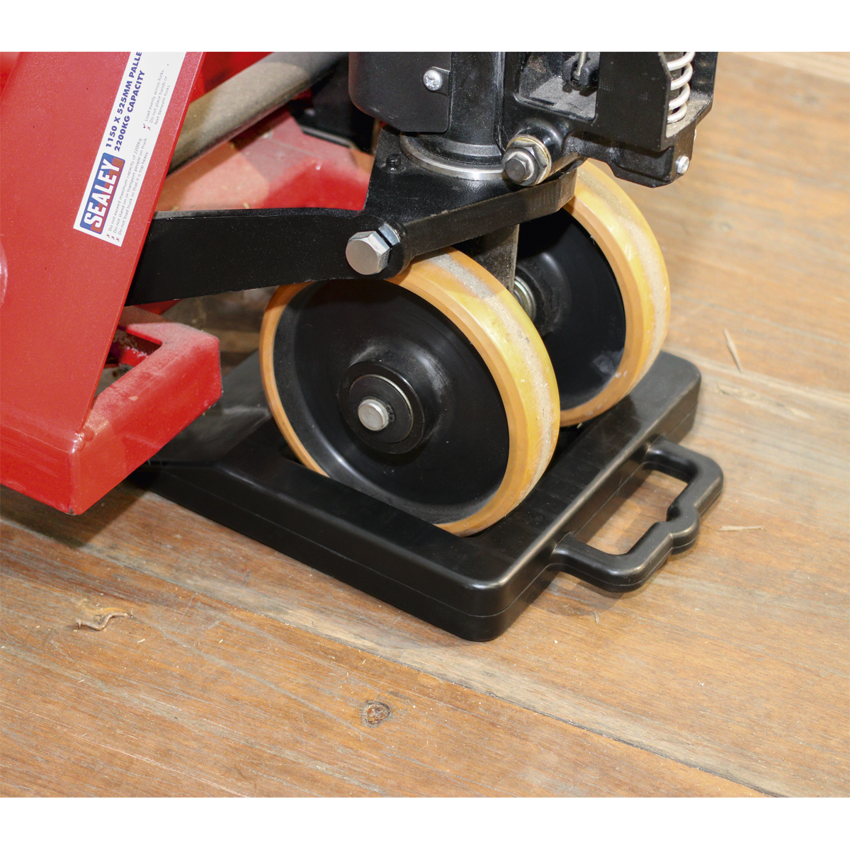 Close-up of the secure wheels on a red industrial jack, positioned on the Sealey Pallet Truck Stop - PTS1, featuring a non-slip durable rubber pad placed on a wooden floor.